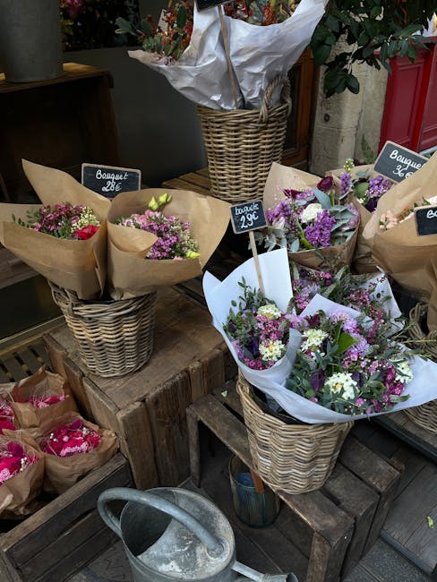A display of floral bouquets arranged in woven wicker baskets, each wrapped in light brown or purple paper, featuring a mix of pink, purple, and white flowers with green foliage. The bouquets are plac