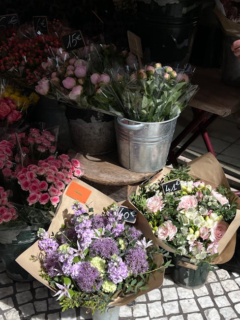 A display of fresh floral bouquets at a florist shop, featuring a variety of flowers arranged in rustic metal and paper-wrapped containers. Prominent arrangements include a bouquet of pale pink roses 