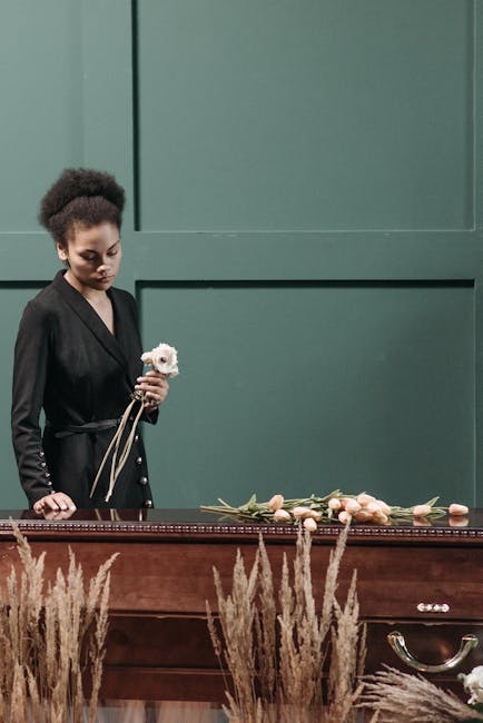 A woman dressed in black, with curly hair styled neatly, is holding a delicate white flower with pale pink accents, complemented by a long cream ribbon. She stands beside a polished wooden table decor