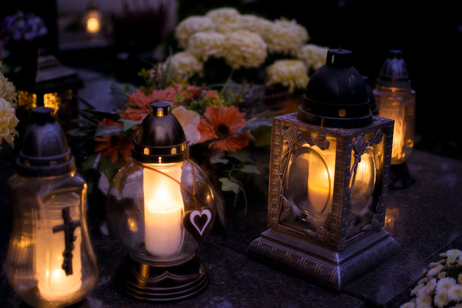 A floral arrangement featuring large white hydrangeas, orange gerbera daisies, and pink roses arranged together on a dark surface, with glass and metal lanterns containing lit candles placed around th