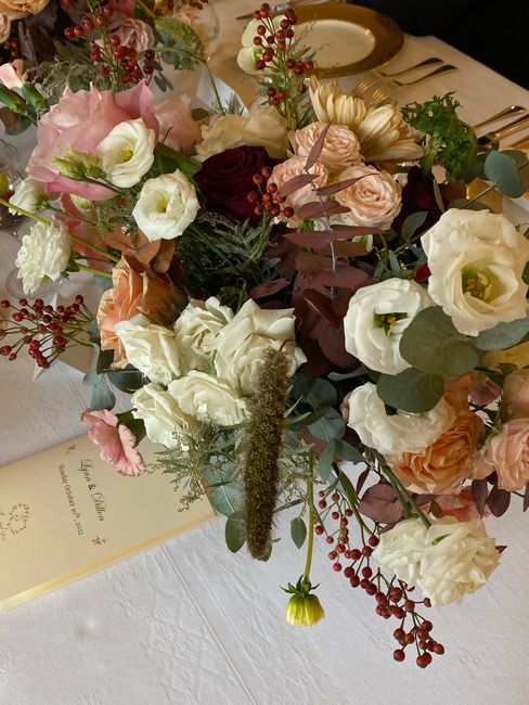 A floral arrangement featuring a mix of roses, eustoma, and lisianthus in shades of blush pink, cream, peach, and burgundy, complemented by sprigs of red berries and lush green foliage including eucal