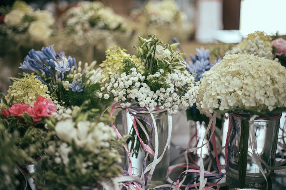 A collection of floral arrangements in clear glass vases featuring predominantly white and pastel-colored flowers, including hydrangeas, roses, and baby's breath, accented with soft pink, blue, and gr